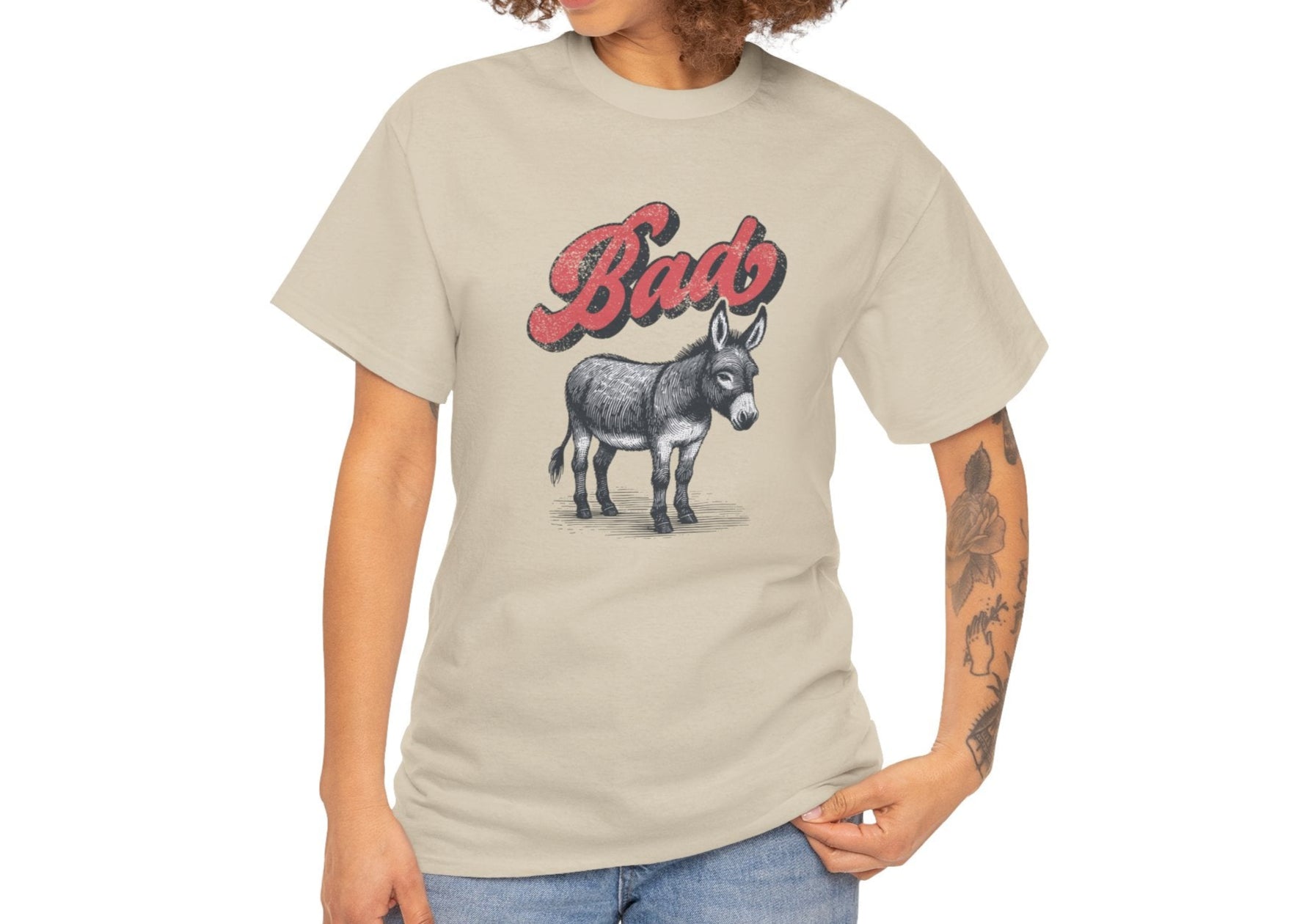 A person wearing a beige T-shirt with the word "Bad" in red text above a detailed donkey illustration, implying "Bad Ass," paired with blue jeans. Part of the Vivid Divergence Sensory Friendly Unisex Tees Range.