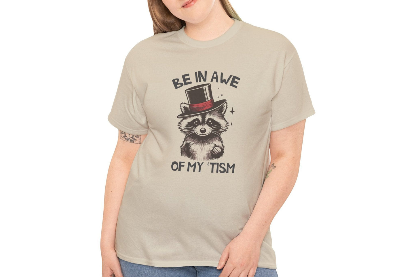 Model wearing a beige T-shirt featuring a raccoon with a top hat and the text "Be In Awe Of My Tism," shown from the front. Part of the Vivid Divergence Sensory Friendly Unisex Tees Range.