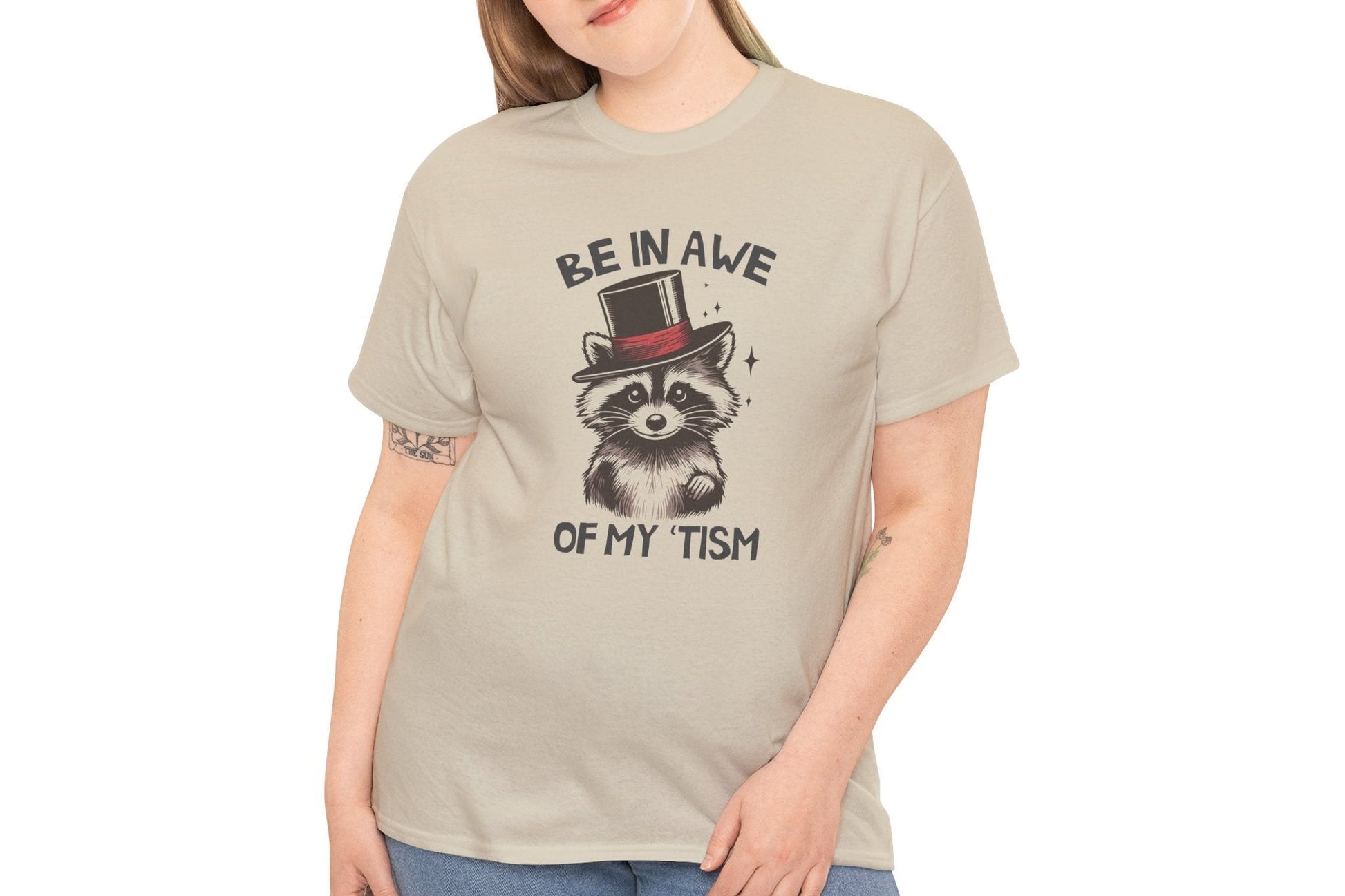 Model wearing a beige T-shirt featuring a raccoon with a top hat and the text "Be In Awe Of My Tism," shown from the front. Part of the Vivid Divergence Sensory Friendly Unisex Tees Range.