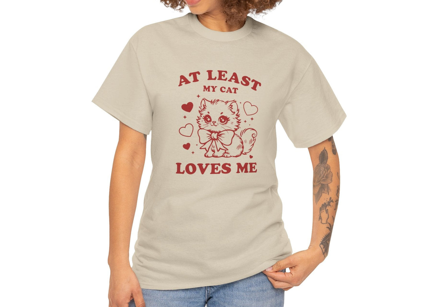 Woman with curly hair wearing a beige T-shirt with red text and cat illustration that reads "At Least My Cat Loves Me." Exclusive release for Valentine’s Day. Part of the Vivid Divergence Sensory Friendly Unisex Tees Range.