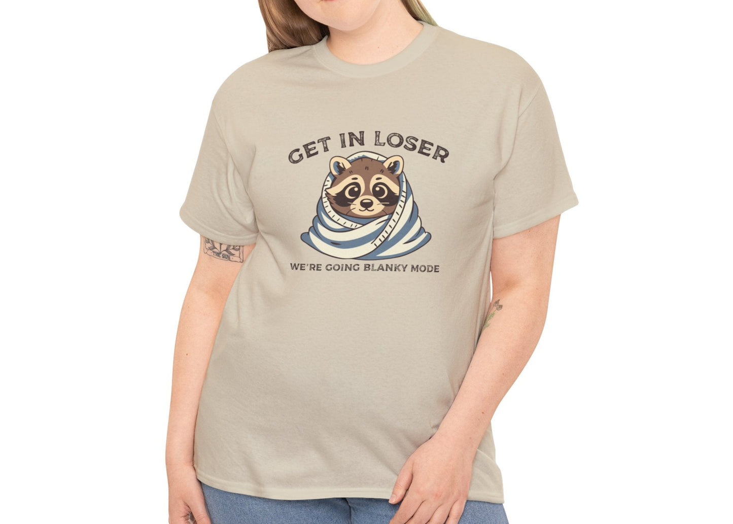 Modeled front view of a beige T-shirt featuring a cute raccoon wrapped in a blanket with text that reads "Get In Loser, We're Going Blanky Mode." Part of the Vivid Divergence Sensory Friendly Unisex Tees Range.