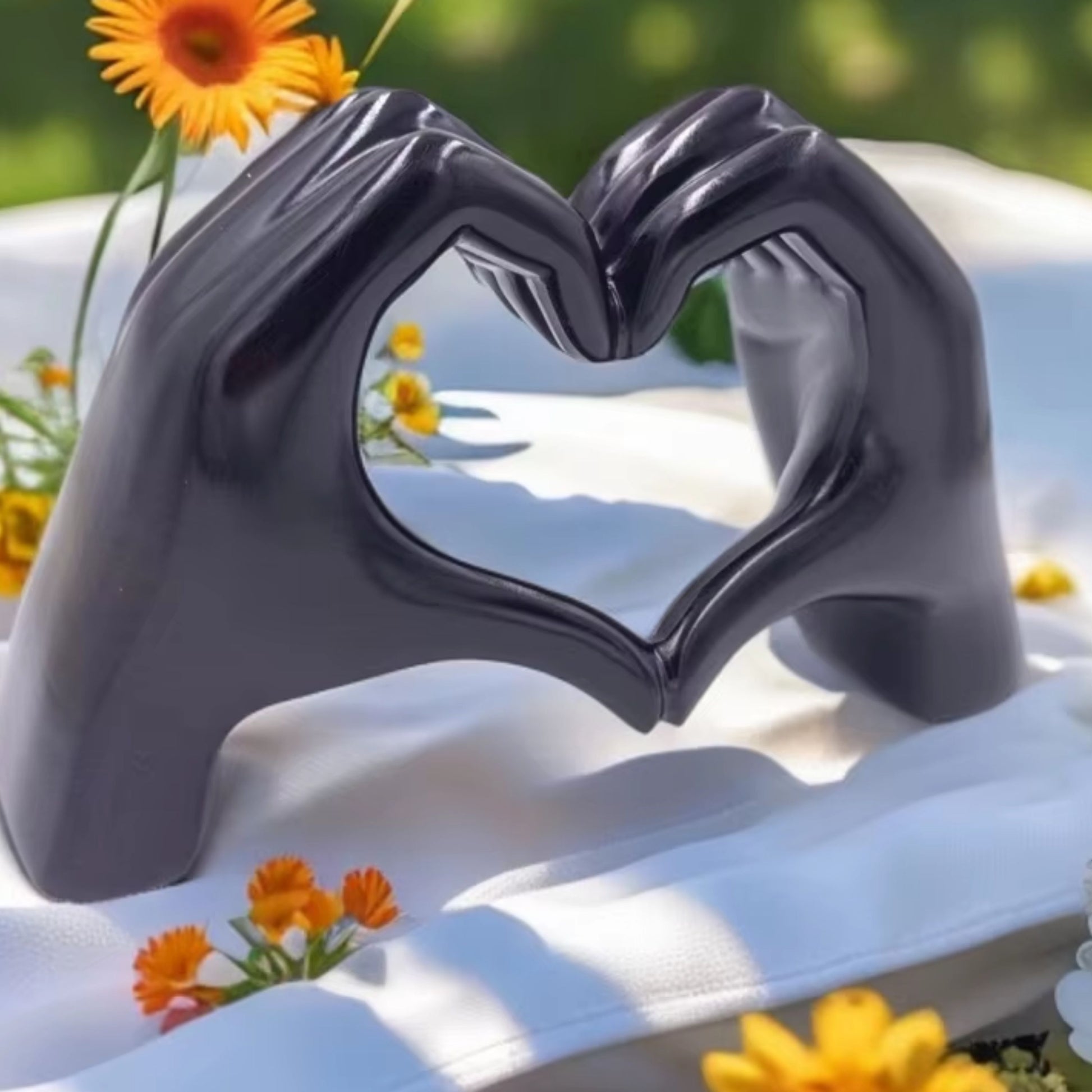 Black glossy hand-shaped sculpture forming a heart, displayed outdoors with flowers. A stylish dopamine decor piece and a great gift for Valentine's Day.