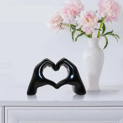 Black glossy hand-shaped sculpture forming a heart on a white table with pink flowers in the background. Perfect dopamine decor and a thoughtful Valentine's Day gift.