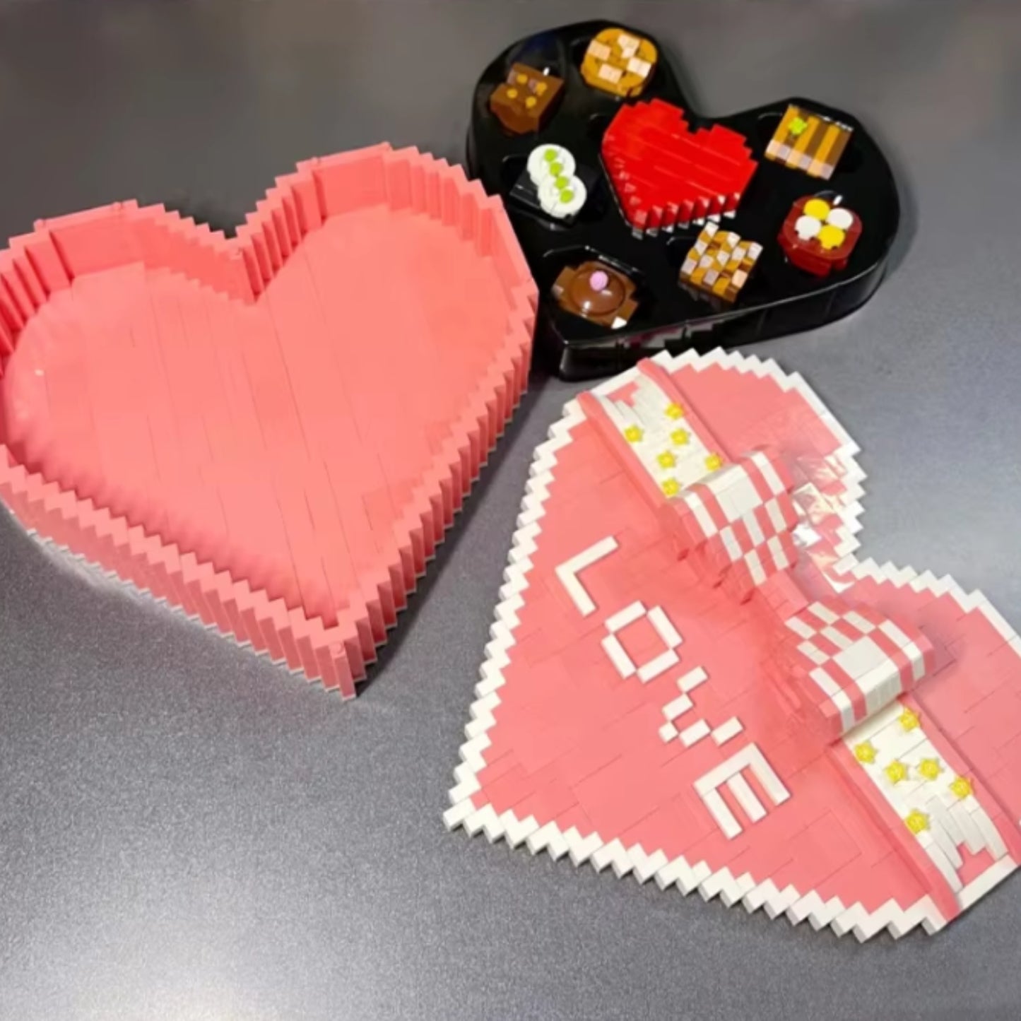 Disassembled pink heart-shaped gift box with lid, chocolates, and black tray made of building blocks.