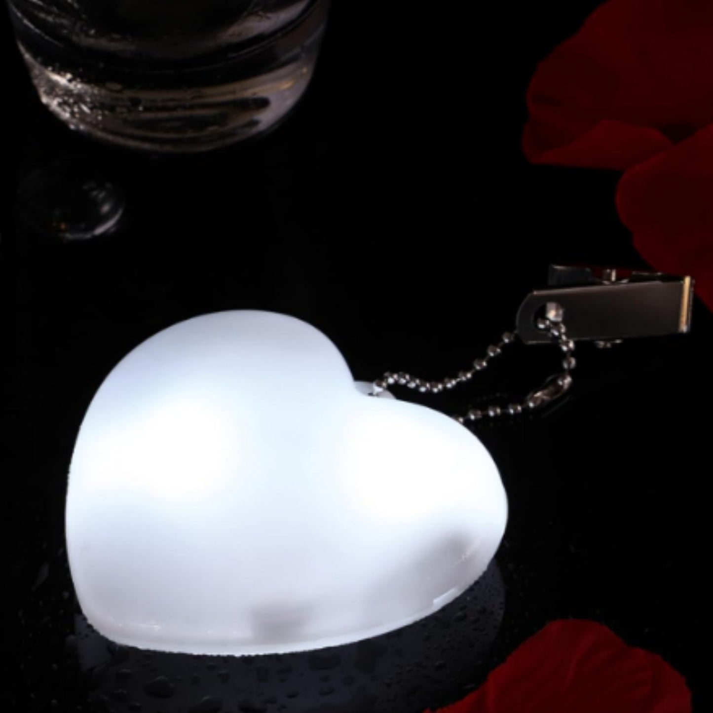 Heart-shaped LED bag clip glowing next to scattered red petals and glass, emphasizing its elegant and versatile design.