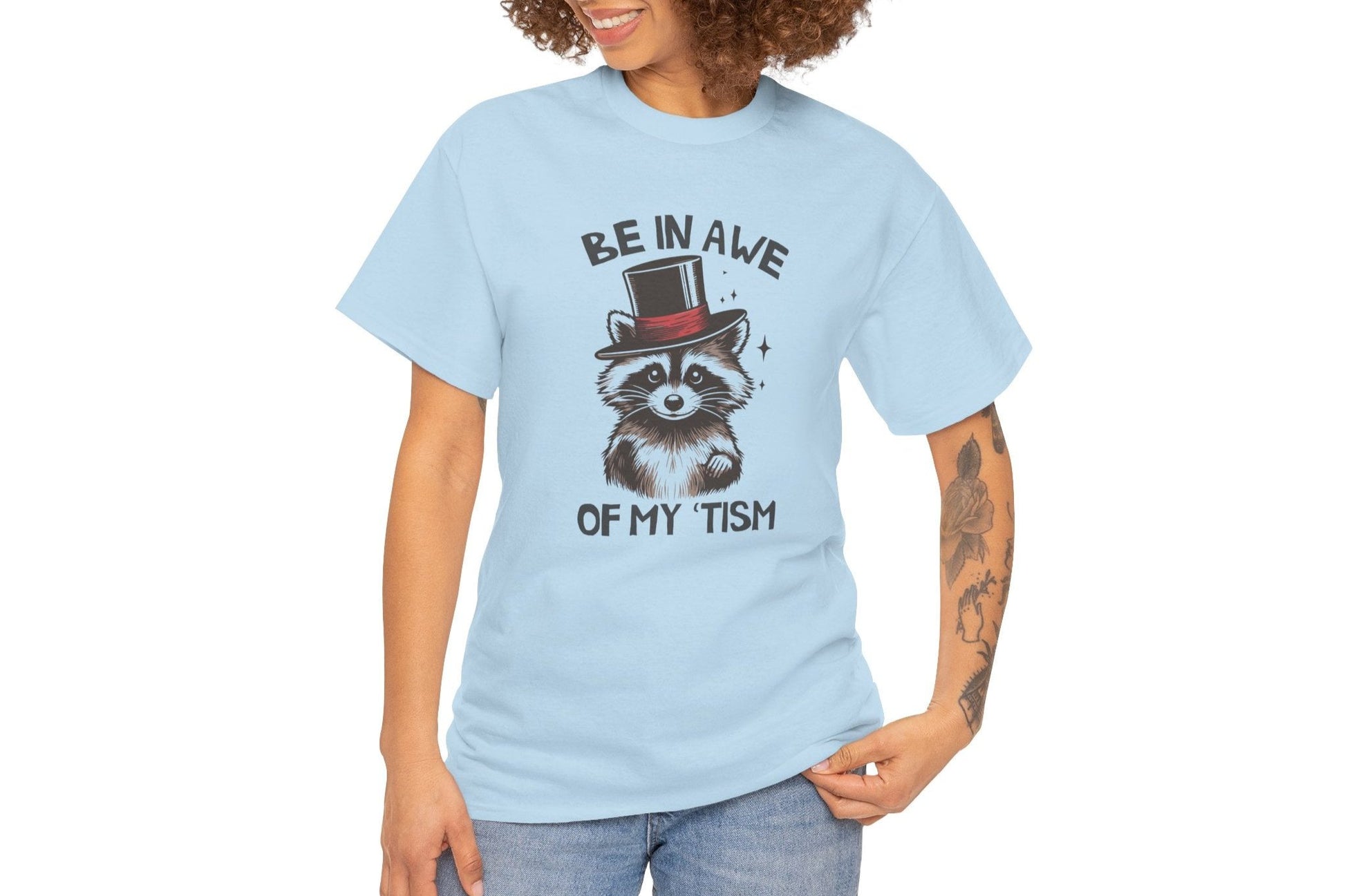 Model wearing a light blue T-shirt featuring a raccoon with a top hat and the text "Be In Awe Of My Tism," shown from the front. Part of the Vivid Divergence Sensory Friendly Unisex Tees Range.