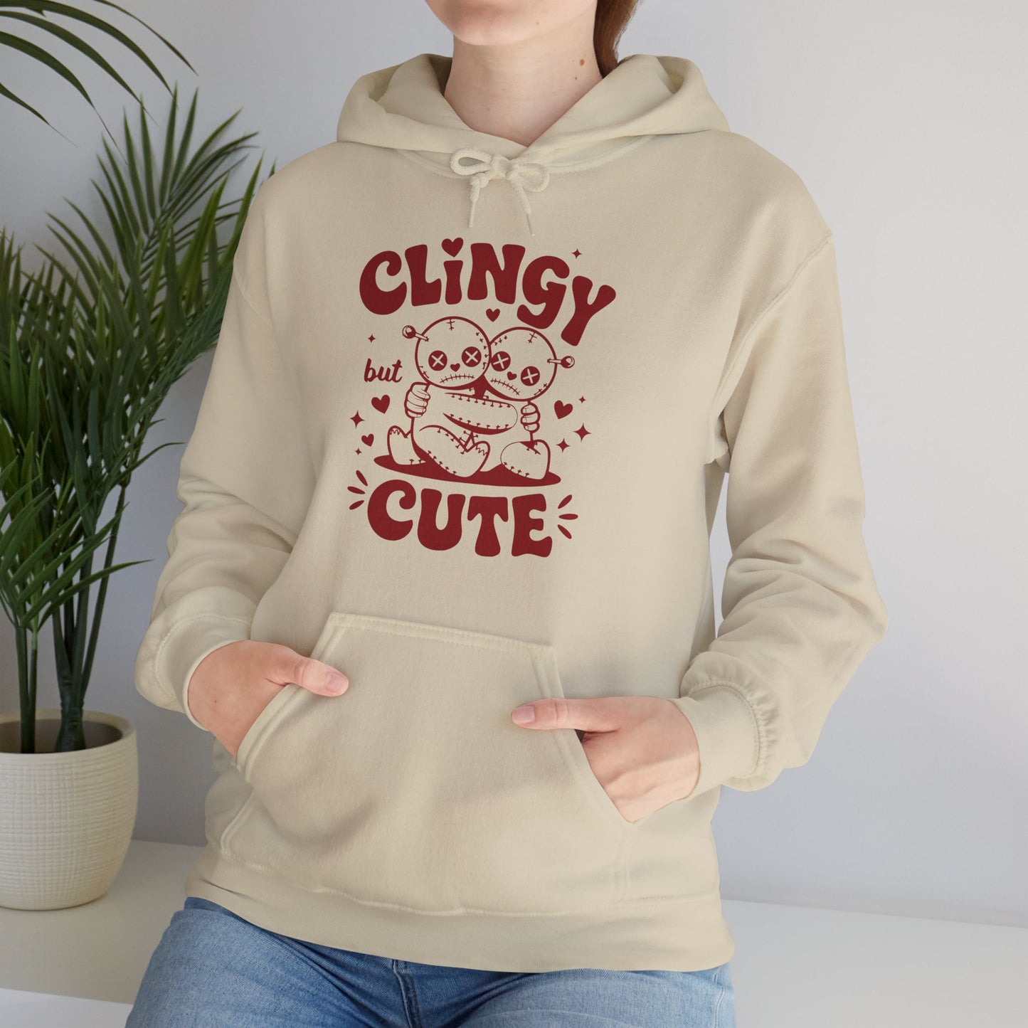 Model wearing a beige hoodie with "Clingy but Cute" text and a cute voodoo doll design in red while seated. Exclusive release for Valentine's Day. Part of the Vivid Divergence Sensory Friendly Sweaters Range.