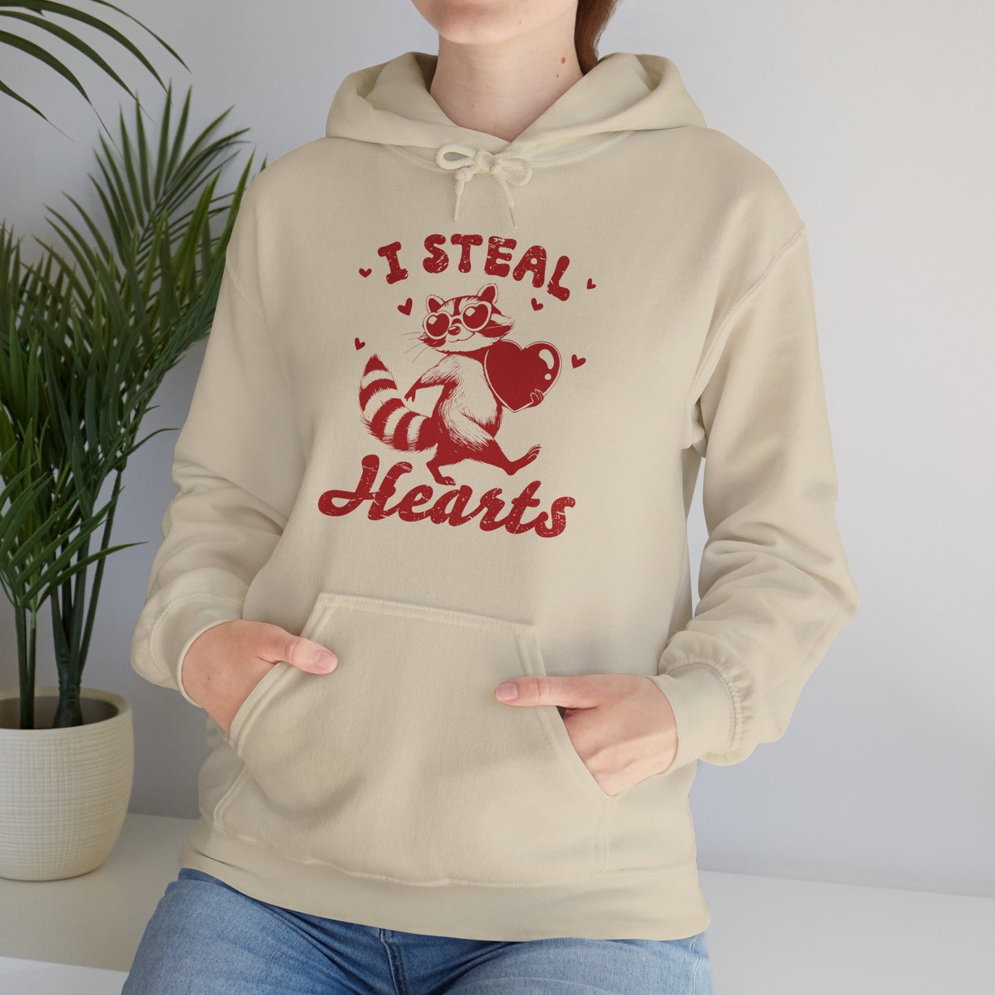 Model wearing a beige hoodie with "I Steal Hearts" text and a cute red Cupid design holding a bow and arrow while seated. Exclusive release for Valentine's Day. Part of the Vivid Divergence Sensory Friendly Sweaters Range.