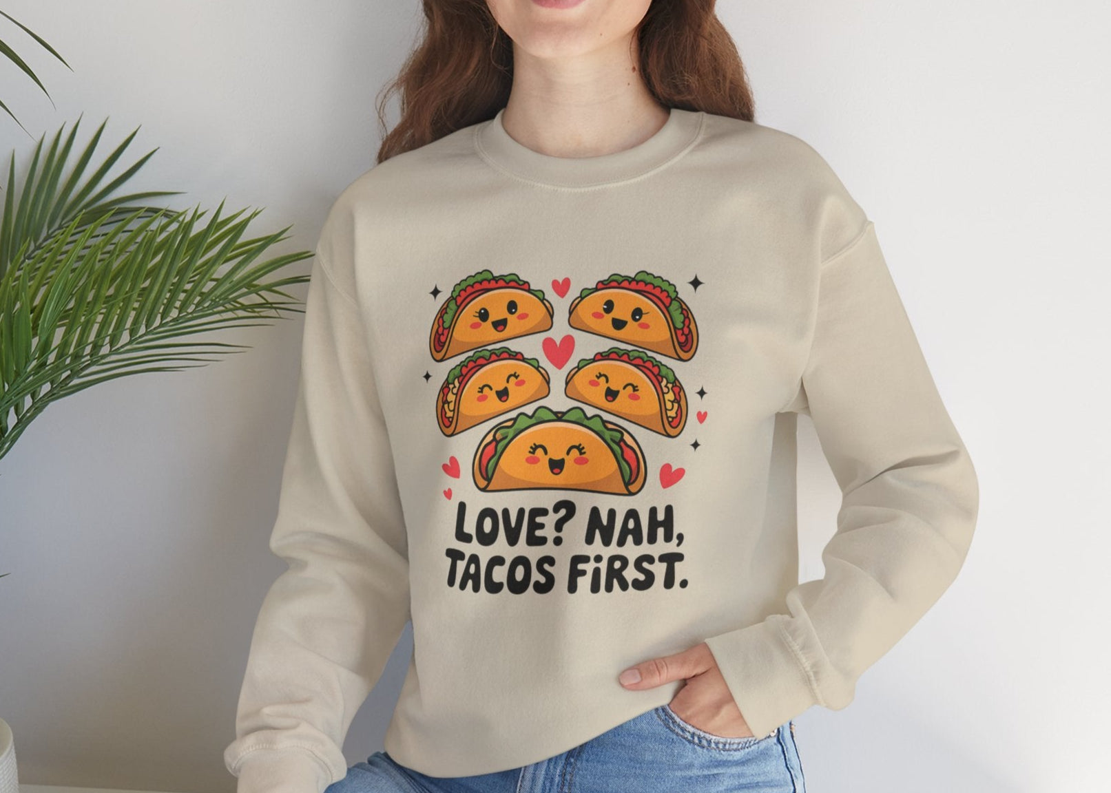 Model wearing a beige sweatshirt with "Love? Nah. Tacos First." text and cute taco characters design in orange and brown while seated. Exclusive release for Valentine's Day. Part of the Vivid Divergence Sensory Friendly Sweaters Range.