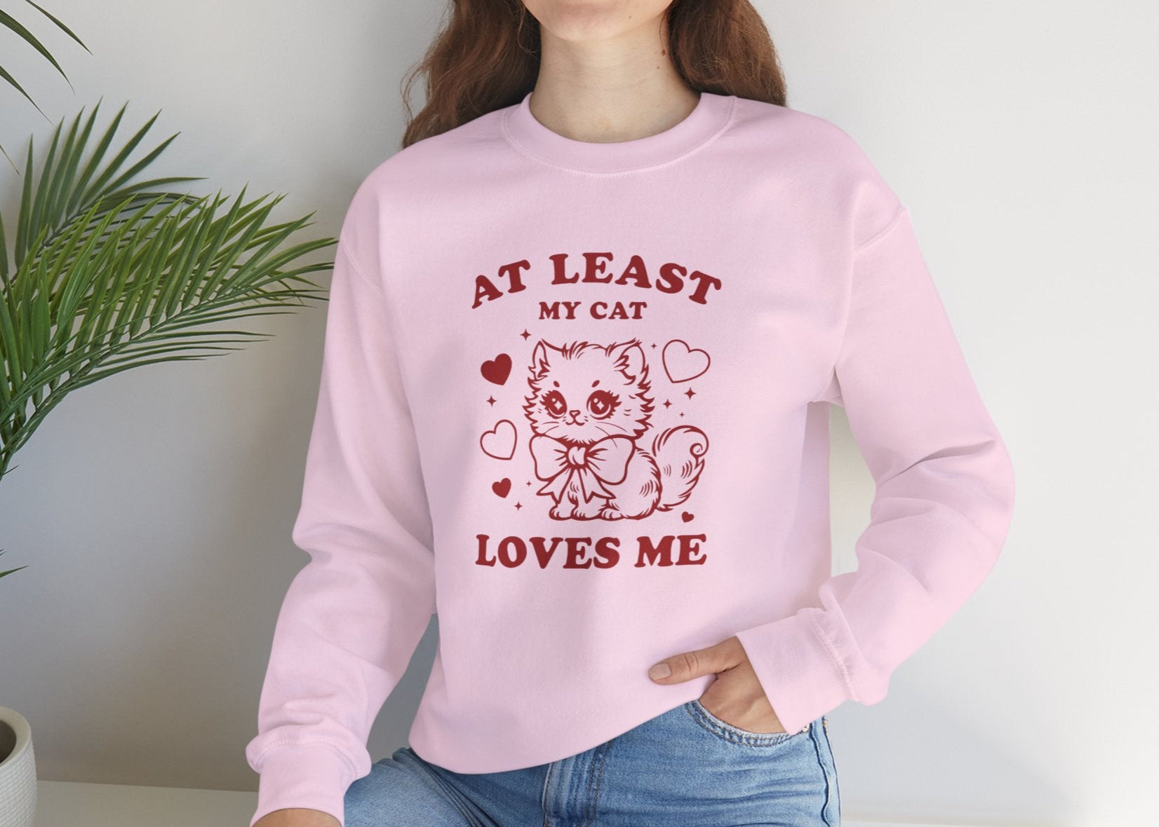 Model wearing a pink sweatshirt with "At Least My Cat Loves Me" text and cartoon cat design in red while seated. Part of the Vivid Divergence Sensory Friendly Sweaters Range.