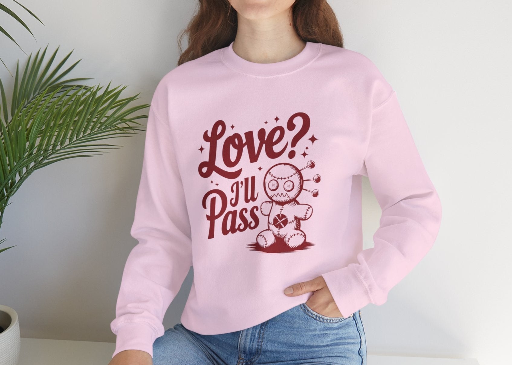 Model wearing a pink sweatshirt with "Love? I'll Pass" text and a cute voodoo doll design in red while seated. Exclusive release for Valentine's Day. Part of the Vivid Divergence Sensory Friendly Sweaters Range.