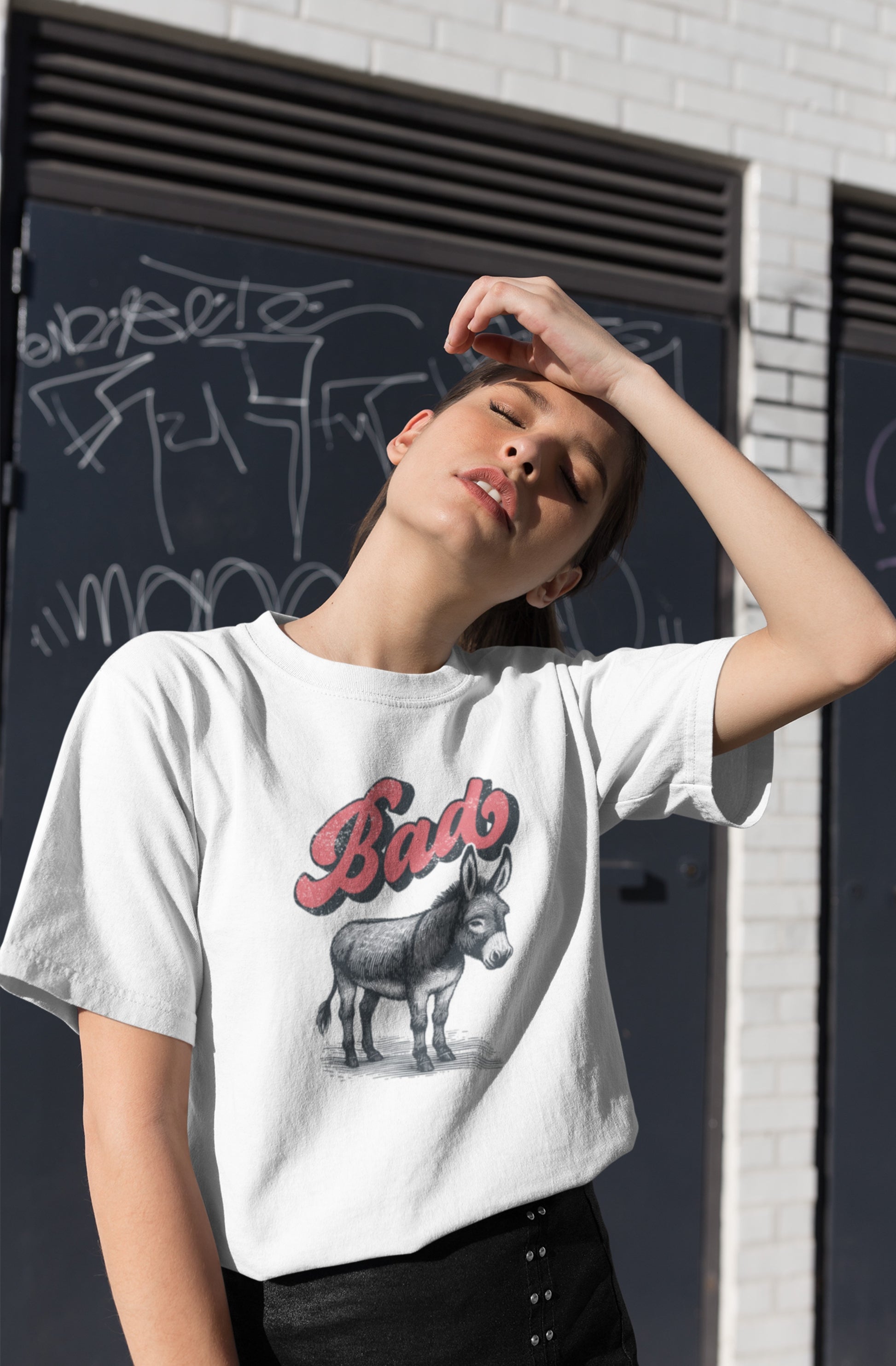 A person wearing a white T-shirt with the word "Bad" in red text above a detailed donkey illustration, implying "Bad Ass," standing against a black chalkboard wall. Part of the Vivid Divergence Sensory Friendly Unisex Tees Range.