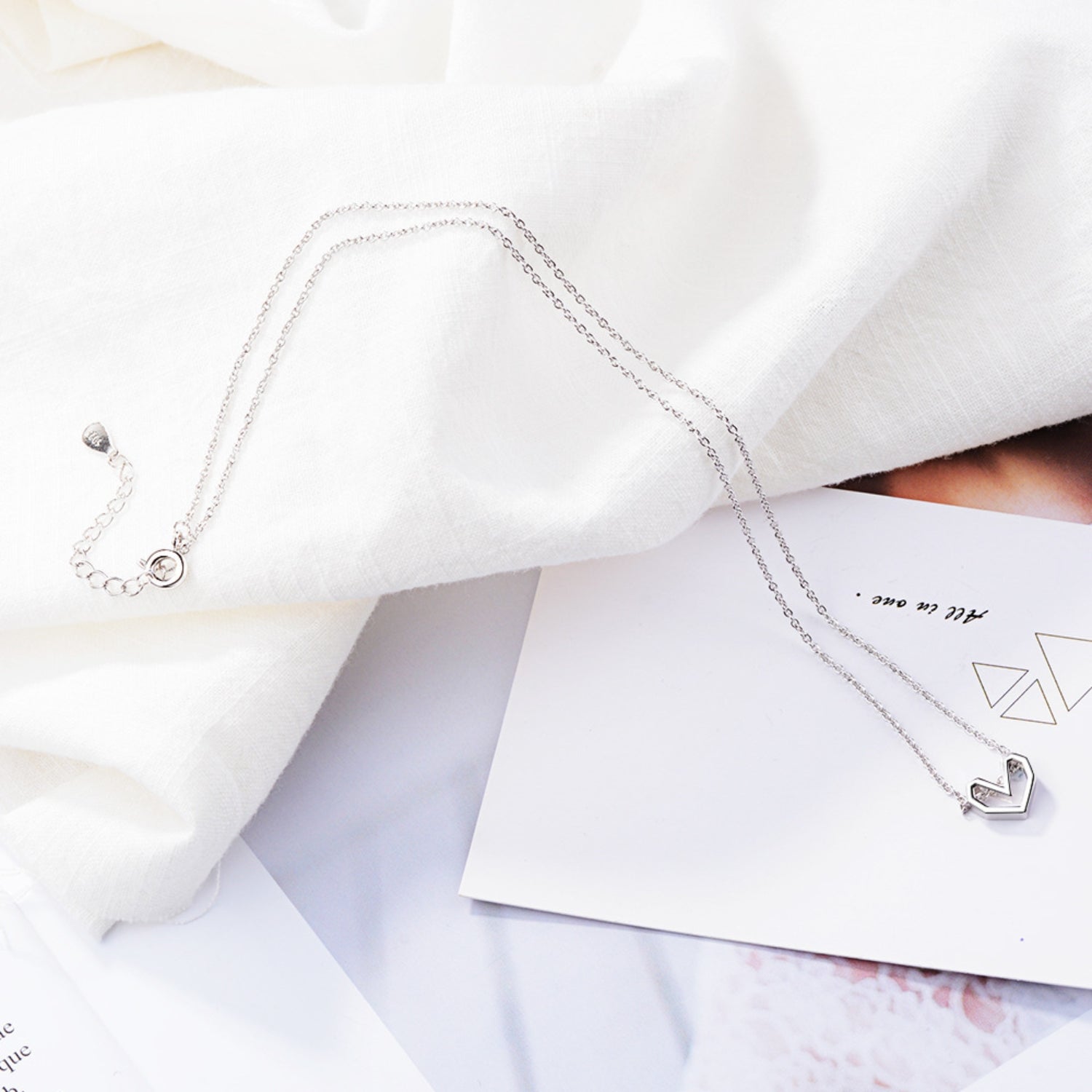 A delicate silver geometric open heart necklace laid out on white fabric with soft lighting, creating a clean and sophisticated aesthetic.