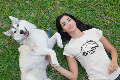 Woman lying on grass with a white dog, wearing a beige T-shirt featuring the text "Excuse Me While I Overthink This." Part of the Vivid Divergence Sensory Friendly Unisex Tees Range.